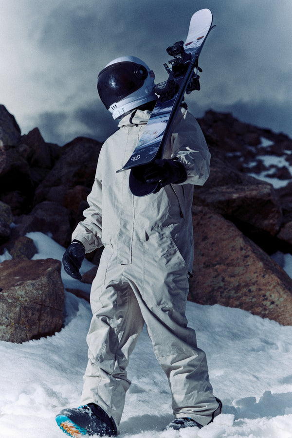 Clad in an astronaut suit, a person carries the Icelantic Skis Nomad 106 on their shoulder while walking across snow-covered, rocky terrain beneath a cloudy sky.