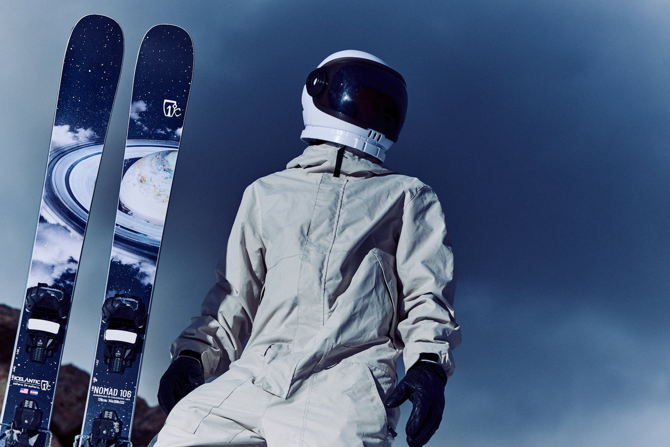 Wearing a white spacesuit and helmet, a person stands next to Icelantic Skis’ Nomad 106 skis, which feature a space-themed design, beneath a cloudy, dark sky.