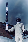 Clad in a white spacesuit and black helmet, a person stands on snowy rocks, holding an upright Icelantic Skis Nomad 106 ski featuring a mountain and moon design, with dark clouds filling the sky behind them.