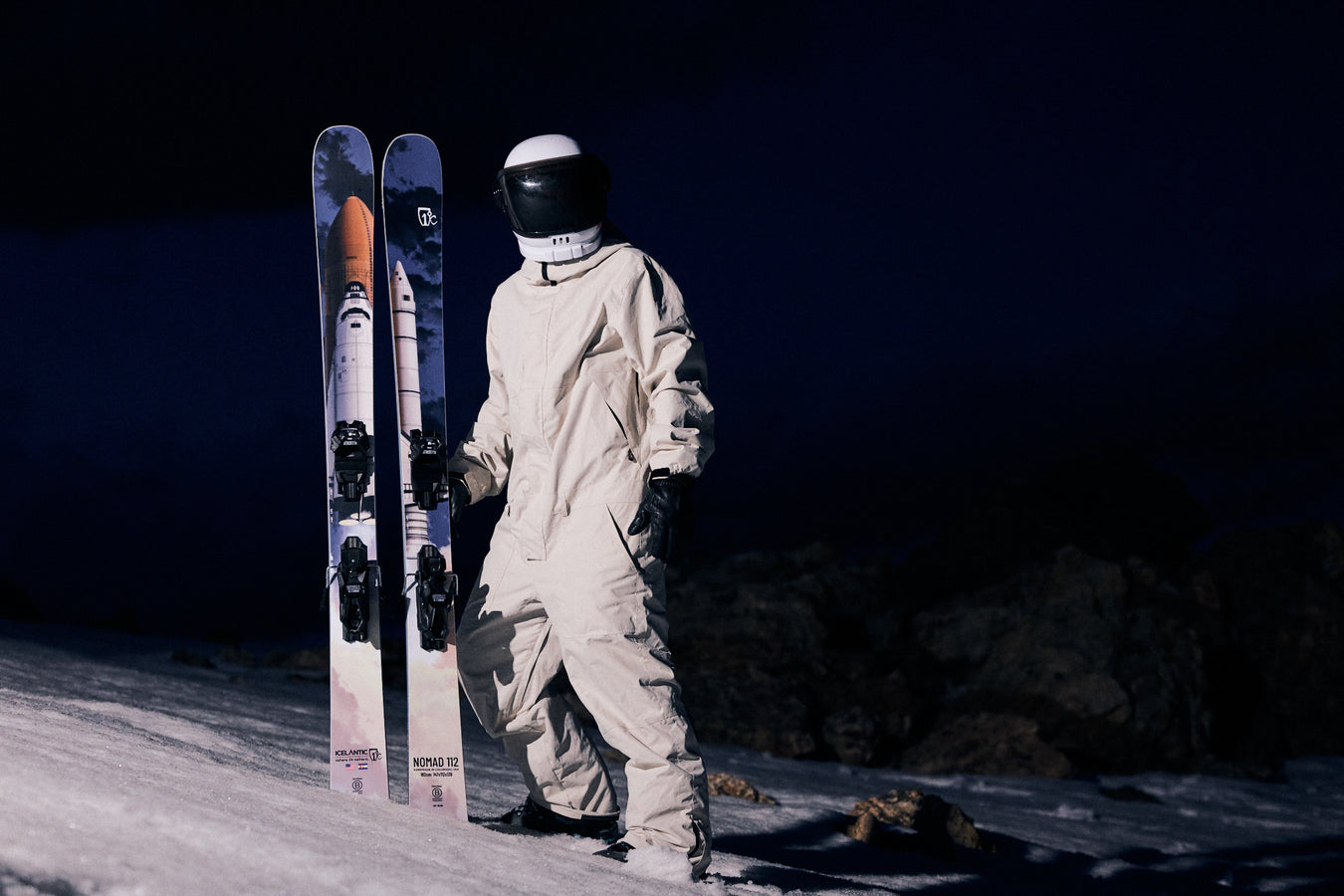 A person in an astronaut suit stands on snow at night, holding Icelantic Skis’ Nomad 112 skis.