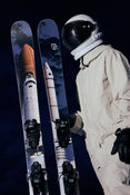 An astronaut in a white suit and helmet holds Icelantic Skis’ Nomad 112 skis, which are decorated with images of a space shuttle, against a dark background.