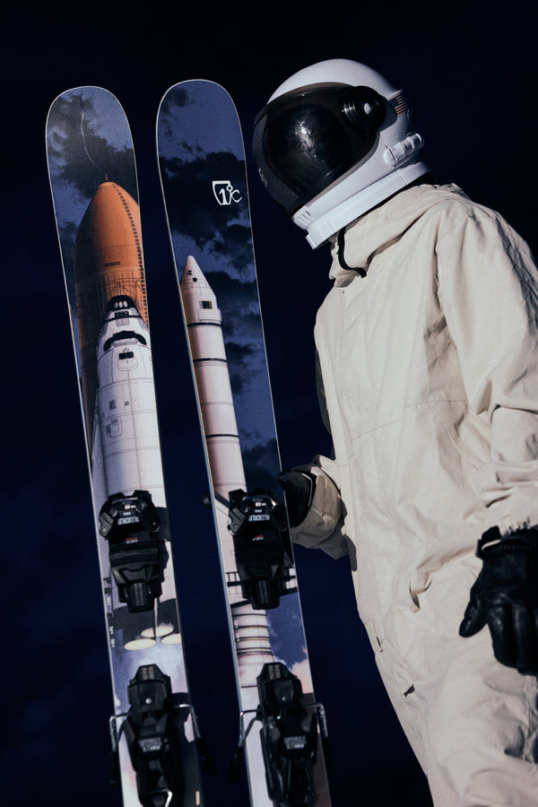 An astronaut in a white suit and helmet holds Icelantic Skis’ Nomad 112 skis, which are decorated with images of a space shuttle, against a dark background.