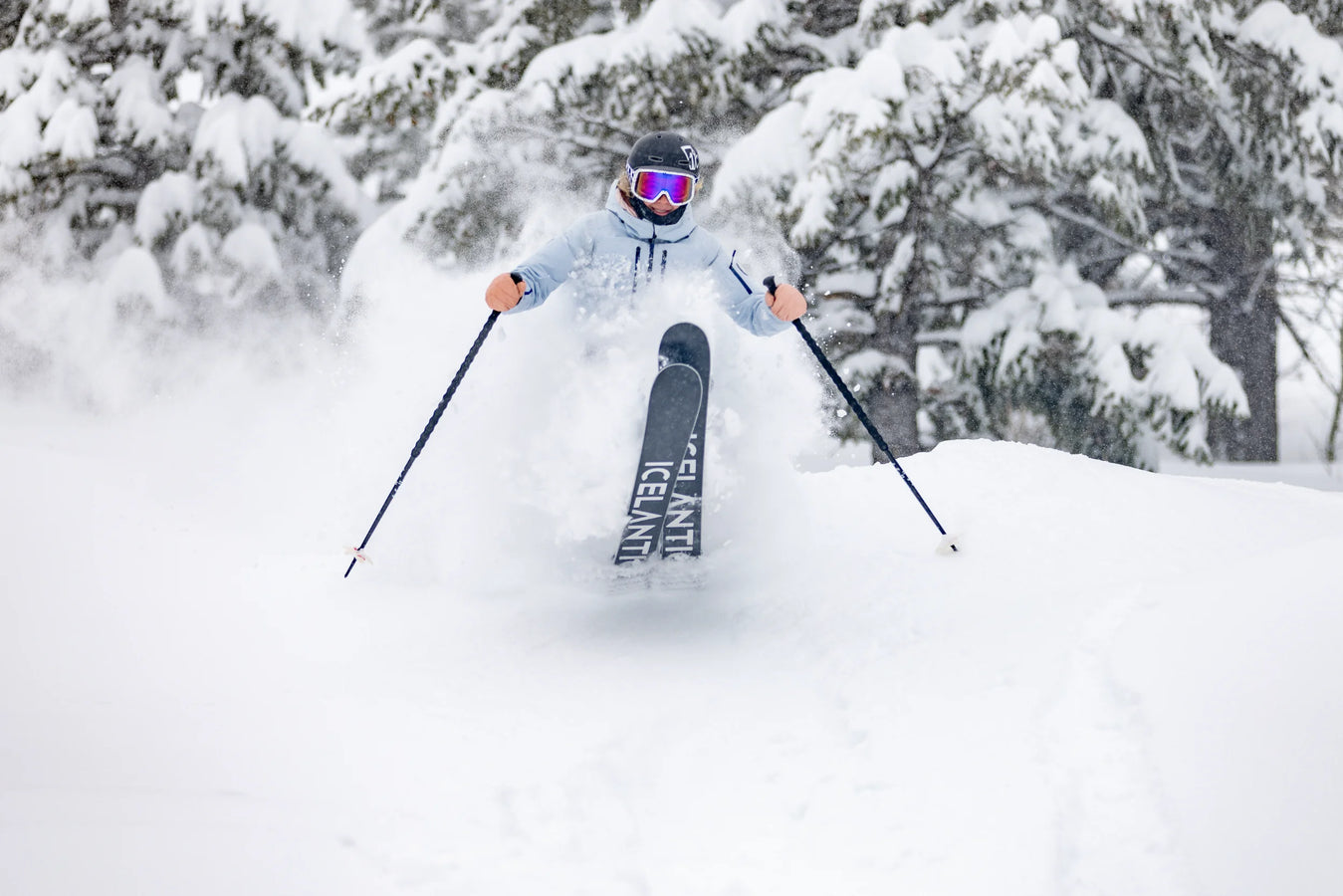 An adventurous skier, equipped with Icelantic Skis’ Maiden 102 and wearing pink goggles, carves through deep powder on a snowy slope lined with snow-covered trees, kicking up fresh snow as they ride.