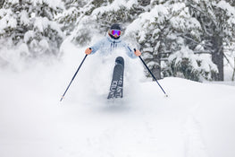 An adventurous skier, equipped with Icelantic Skis’ Maiden 102 and wearing pink goggles, carves through deep powder on a snowy slope lined with snow-covered trees, kicking up fresh snow as they ride.