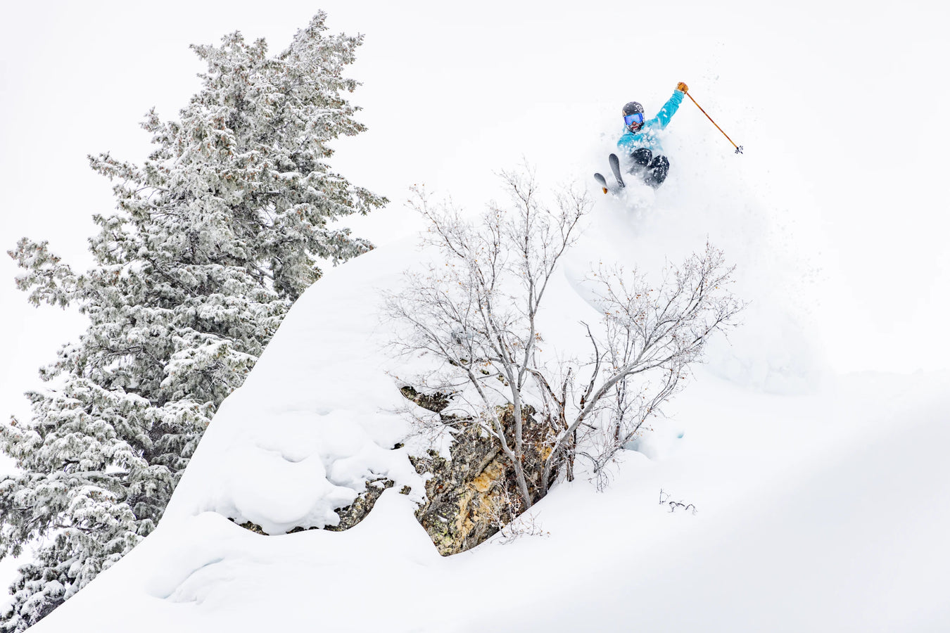 Wearing blue gear, a skier launches off a snow-covered cliff surrounded by trees and fresh powder, riding on Icelantic Skis’ Nomad 106 against a white winter backdrop.