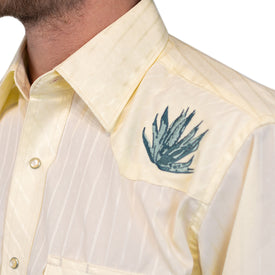 A close-up of a person wearing the Wrangler Agave Pearl Snap Shirt by Icelantic Skis, a light yellow cowboy dress shirt adorned with blue-green agave plant embroidery on the left shoulder. The background is white.