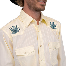 A man wearing a light yellow Wrangler Agave Pearl Snap Shirt by Icelantic Skis—a cowboy dress shirt featuring embroidered teal agave plants on the shoulders and two buttoned chest pockets—perfectly captures the classic western clothing style.