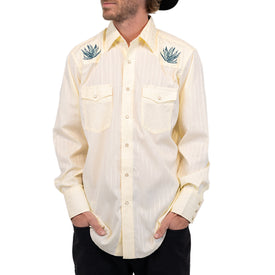 A man stands with his hands in his pockets, looking to the side, dressed in the Icelantic Skis Wrangler Agave Pearl Snap Shirt—a cream-colored, long-sleeve western shirt featuring subtle stripes, two chest pockets, and blue agave plant embroidery on the shoulders.
