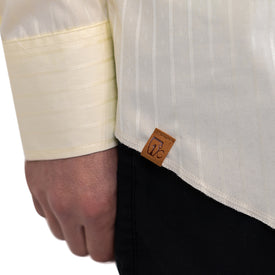 A close-up of a person wearing the Wrangler Agave Pearl Snap Shirt by Icelantic Skis, a cream-colored, long-sleeve cowboy dress shirt with a subtle pattern, paired with black pants and featuring a brown tag with a small logo on the hem.