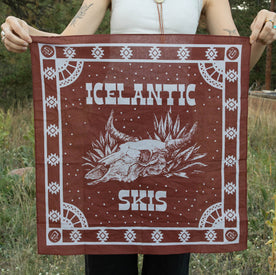 A person holds the Agave Skull Bandana by Icelantic Skis, made in Denver from lightweight cotton. The brown bandana features the “Icelantic Skis” text and a central animal skull surrounded by plants, standing out against a grassy outdoor background.
