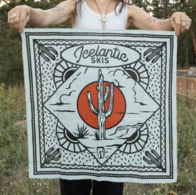 A person holds the Cactus Bandana by Icelantic Skis, a square, lightweight cotton bandana made in Denver. The design features a desert scene with a cactus in front of a red sun, accented in black, white, and orange. "Icelantic Skis" is printed at the top.