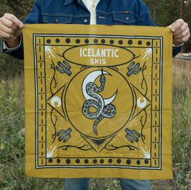 A person holds the Rattlesnake Bandana by Icelantic Skis—a gold cotton accessory featuring a snake design, crescent moons, skis, and "ICELANTIC SKIS" printed at the center. Made in Denver from lightweight cotton, this bandana stands out against an outdoor backdrop of grass and trees.