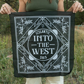 Outdoors, a person holds the Icelantic Skis Into The West Bandana—a lightweight black cotton accessory featuring bold white text and floral corner designs, expertly crafted in Denver.