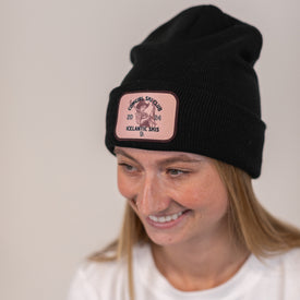 A smiling person wearing the Icelantic Skis Cowgirl Ski Club Beanie in black, featuring a tight knit and a custom pink patch with an illustrated cat and the text "General Shulko 2018 Icelandic Skis.