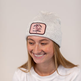 A woman with straight blonde hair smiles while wearing the Cowgirl Ski Club Beanie in White Heather from Icelantic Skis, which features a custom patch. She is dressed in a plain white shirt.
