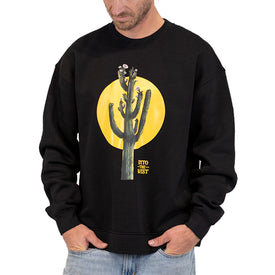 A man is wearing the Icelantic Skis Cactus Crew, a black unisex crewneck sweatshirt featuring a graphic of a cactus set against a large yellow sun with the words INTO THE WEST. Crafted from recycled polyester fleece, this cozy Nomad 106 crew style has him standing with his hands in his pockets, looking down.