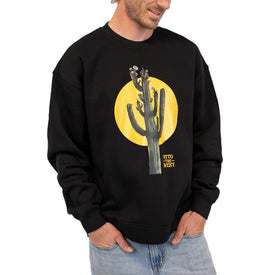 A man in the Icelantic Skis Cactus Crew—a black unisex crewneck sweatshirt featuring a cactus and yellow sun graphic—stands with his hands in his jeans pockets, smiling and glancing slightly to the side. Crafted from recycled polyester fleece, this sweatshirt offers both comfort and style.