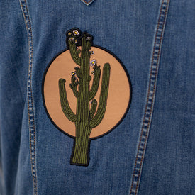The Wrangler Cactus Denim Jacket - Women's by Icelantic Skis showcases a round patch depicting a green cactus with delicate flowers on a tan background, offering a striking tribute to classic western style.