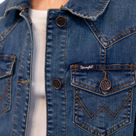 Close-up of a person wearing the Icelantic Skis Wrangler Cactus Denim Jacket for women, featuring blue denim with brown stitching, metal buttons, and a small logo patch above the left chest pocket—offering a contemporary twist on classic western style. The person is also wearing a white shirt underneath.