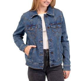 A woman wearing the Icelantic Skis Wrangler Cactus Denim Jacket over a white shirt and black jeans stands with one hand in her jacket pocket, smiling against a white background.