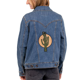 A woman stands smiling, facing away from the camera and wearing the Icelantic Skis Wrangler Cactus Denim Jacket, which features a green cactus patch inside a beige circle on the back—a stylish choice for those who appreciate western fashion with a modern flair.
