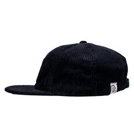 The Icelantic Skis Cowgirl Ski Club Corduroy Cap - Black is a stylish six-panel snapback hat with a flat brim, shown here in a side view. It features classic black corduroy fabric and an adjustable back strap, accented by a small white rectangular patch bearing the minimalist Cowgirl Ski Club logo.