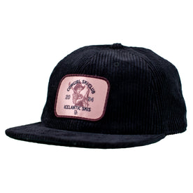 The Cowgirl Ski Club Corduroy Cap - Black by Icelantic Skis is a classic six-panel snapback hat, designed in black corduroy, featuring a pink patch that reads "Cowgirl Ski Club 2024 Icelantic Skis" and displays an image of a person skiing in a cowboy hat.