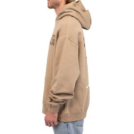 A person wearing a tan Cowboy Up Hoodie by Icelantic Skis, made from recycled polyester, along with light blue jeans, is standing in profile facing left. The hoodie has text on the front that is mostly obscured. The background is plain white.