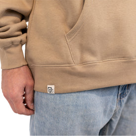 A person wearing a tan Cowboy Up Hoodie by Icelantic Skis and light blue jeans stands with their hand by their side. A small white logo tag is visible on the unisex hoodie near the pocket, highlighting its modern design crafted from recycled polyester.