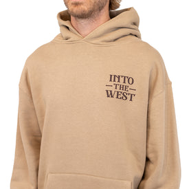 A person wearing a tan unisex hoodie—the Cowboy Up Hoodie by Icelantic Skis—made from recycled polyester, with the words INTO THE WEST printed in black on the upper left chest. The image only shows the person from the shoulders to mid-torso.