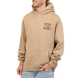 Man wearing a tan Icelantic Skis Cowboy Up Hoodie made from recycled polyester, featuring the text INTO THE WEST printed on the left chest, standing with hands partially in the front pocket, and dressed in light blue jeans.