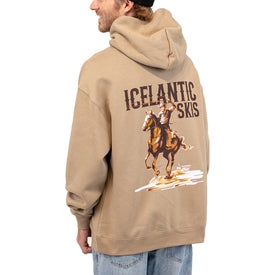 A man stands sideways and smiles, wearing the unisex Cowboy Up Hoodie by Icelantic Skis—a tan Nomad 100 hoodie made from recycled polyester, featuring “ICELANTIC SKIS” and an illustration of a cowboy riding a horse on the back.