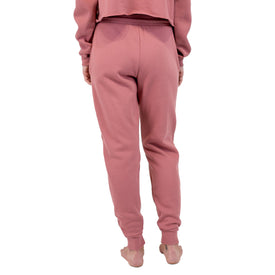 A person stands barefoot, facing away, wearing pink Cowgirl Ski Club Joggers and a matching cropped sweatshirt from Icelantic Skis, isolated on a white background.