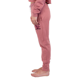 A person stands barefoot in a side profile view, wearing the Cowgirl Ski Club Joggers by Icelantic Skis with a matching pink cropped sweatshirt, set against a white background.