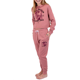 A smiling person wearing Icelantic Skis' Cowgirl Ski Club Joggers with matching pink fleece and maroon graphics, standing barefoot against a white background.