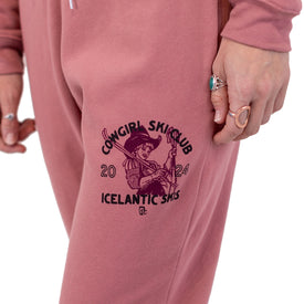 A person wearing pink Cowgirl Ski Club Joggers from Icelantic Skis, featuring a cowgirl graphic on the leg and "COWGIRL SKI CLUB 2024" text. The person is also wearing rings on their fingers.