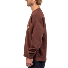 A man with light brown hair stands in profile facing left against a white background, wearing the Icelantic Skis Howdy Crew unisex brown pullover sweatshirt and black pants.