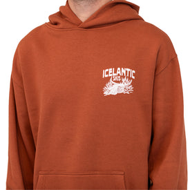 Person wearing a rust-colored unisex oversized Mezcal Skull Hoodie by Icelantic Skis, featuring a white logo and mountain design on the left chest. Only the upper chest and neck are visible, offering a glimpse of this sustainable hoodie crafted from recycled polyester for comfortable wear.