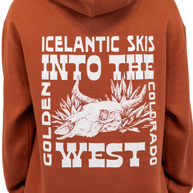 A person wearing the Icelantic Skis Mezcal Skull Hoodie, a rust-colored unisex oversized hoodie featuring white graphic text—"Icelantic Skis Into the West Golden Colorado"—and an illustration of a cow skull with agave plants.
