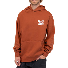 A person wearing the Icelantic Skis Mezcal Skull Hoodie—a unisex, oversized orange-brown hoodie featuring a white Icelantic Skis logo and graphic on the chest—stands against a white background.