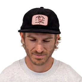 A man with light brown hair and a beard wears the Icelantic Skis Cowgirl Ski Club Corduroy Cap in black—a six-panel hat featuring a pink patch—paired with a light gray sweatshirt, looking down against a white background.