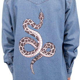 A person wearing a blue denim jacket featuring a striking black and white rattlesnake illustration on the back, reminiscent of the bold Wrangler Rattlesnake Denim Shirt by Icelantic Skis.