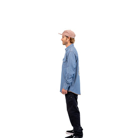 A man stands in profile facing left, wearing a pink cap, a Wrangler Rattlesnake Denim Shirt by Icelantic Skis styled as a western work shirt, black pants, and black shoes, against a plain white background.