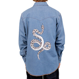 A person stands with their back to the camera, wearing the Icelantic Skis Wrangler Rattlesnake Denim Shirt in light blue, featuring a bold black and white Nomad 94 snake design printed on the back.