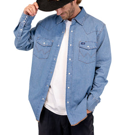 A man wearing a cowboy hat, a light blue Wrangler Rattlesnake Denim Shirt by Icelantic Skis, and a white T-shirt looks down while holding the brim of his hat.