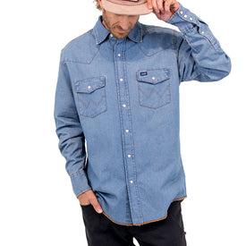 A person wearing an Icelantic Skis Wrangler Rattlesnake Denim Shirt in light blue, black pants, and a tan cap, looking down while holding the brim of the cap with one hand.