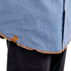 A close-up of a person wearing the Wrangler Rattlesnake Denim Shirt by Icelantic Skis, featuring blue denim with a frayed hem and brown stitching, paired with black pants. A small brown tag is sewn onto the hem, showcasing the Nomad 94 snake design for an authentic western work shirt vibe.