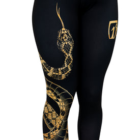 Introducing the Rattlesnake Leggings by Icelantic Skis—sleek black leggings showcasing an intricate gold snake illustration winding up one leg, complemented by a gold geometric logo on the opposite thigh. Featuring a flattering high-waisted waistband, these leggings are crafted from Hyper Wiking WIKMAX Fabric to deliver exceptional comfort and performance.