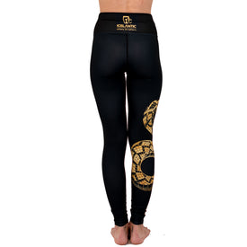 A person stands barefoot, facing away, wearing high-waisted black Rattlesnake Leggings by Icelantic Skis, featuring gold geometric patterns and Icelantic branding on the waistband. Made from Hyper Wiking WIKMAX Fabric, these leggings combine style and performance against a clean white background.