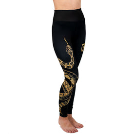 A person stands barefoot against a white background, wearing Icelantic Skis' Rattlesnake Leggings—black high-waisted leggings featuring a gold snake design along the leg.